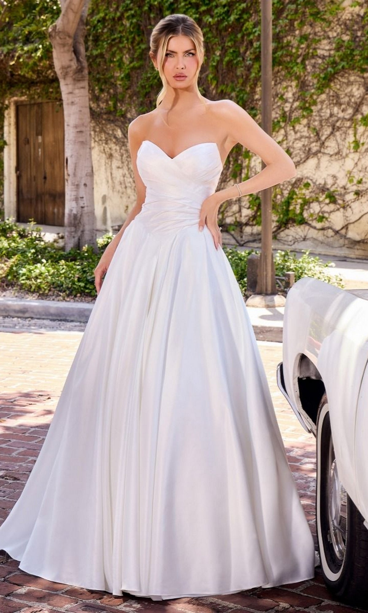 A woman wearing the Ladivine WN015 A-Line Long Classic White Dress in off-white, featuring a strapless sweetheart neckline, stands outdoors beside a white car with greenery and brick paving in the background.